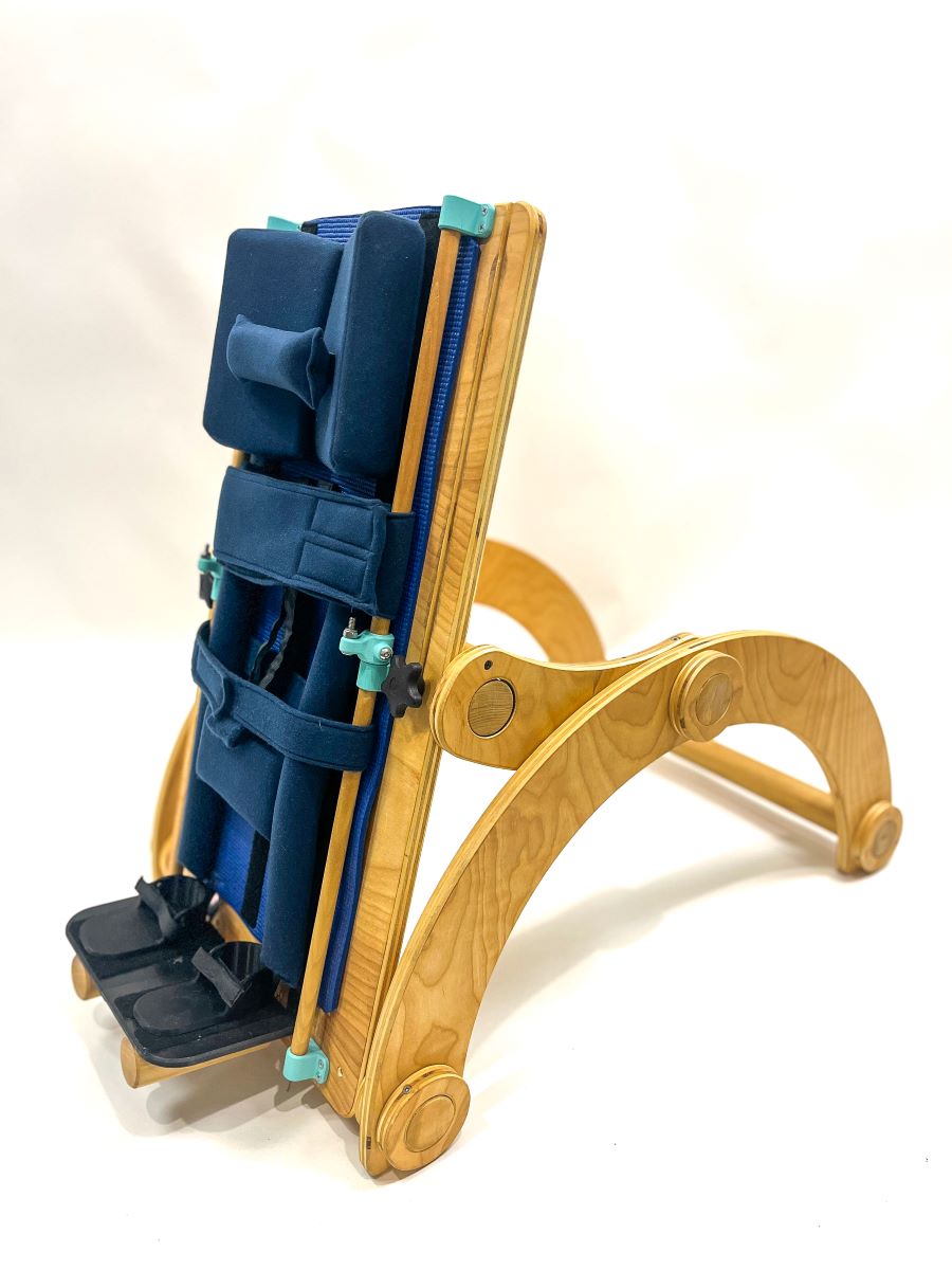 Wood and cardboard children’s standing support device, featuring adjustable back and curved legs, a padded blue seat and head pillows, footrests, and adjustable Velcro harness.