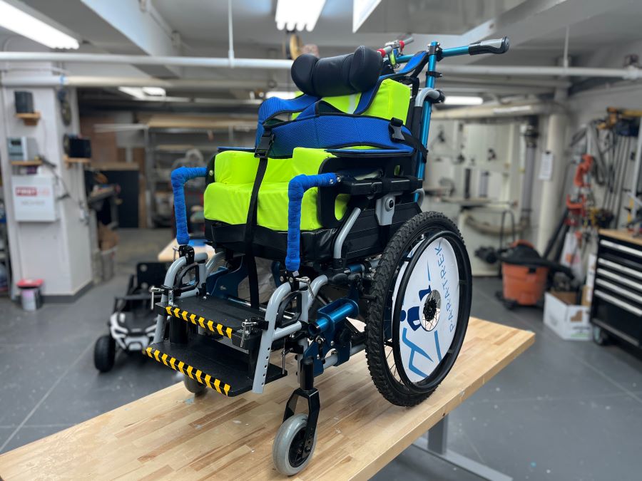 Wheelchair adapted with bright green seat cushions, blue support straps, and large rear wheels, shown on a wooden workbench in a workshop.