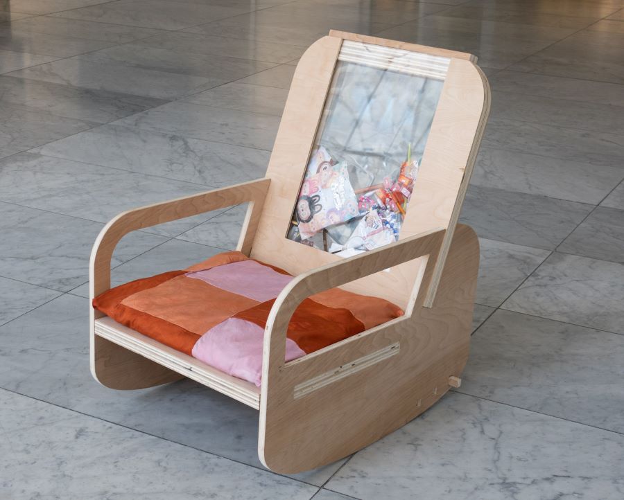 A rocking chair made of light plywood with an orange and pink patchwork cushion. The backrest has a transparent panel filled with plastic packaging materials, highlighting consumption and recycling.