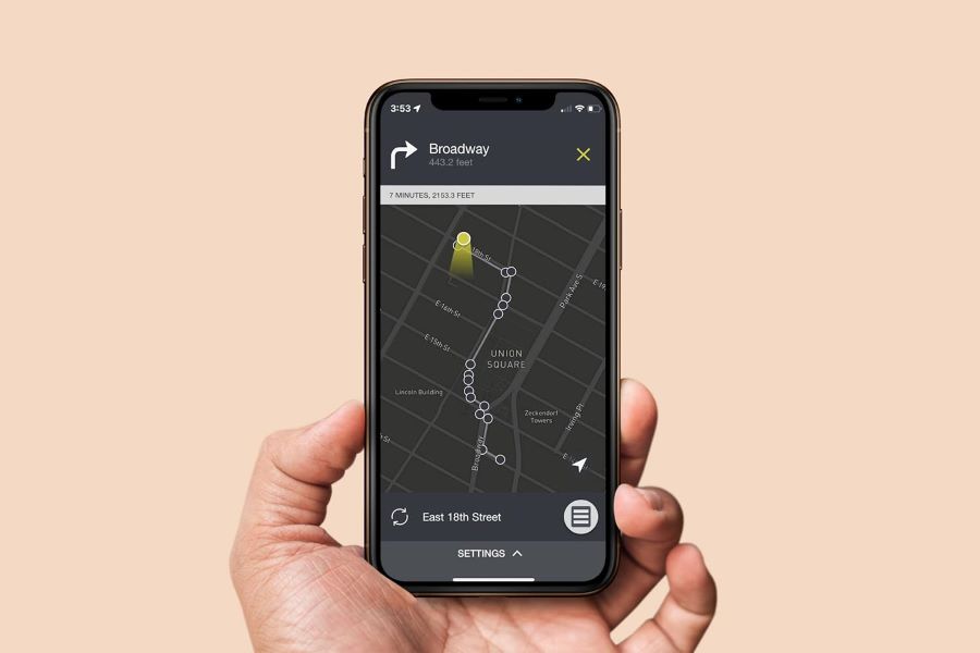 A hand holding a smartphone displaying a navigation app with a highlighted walking route on a dark map background, showing step-by-step directions.