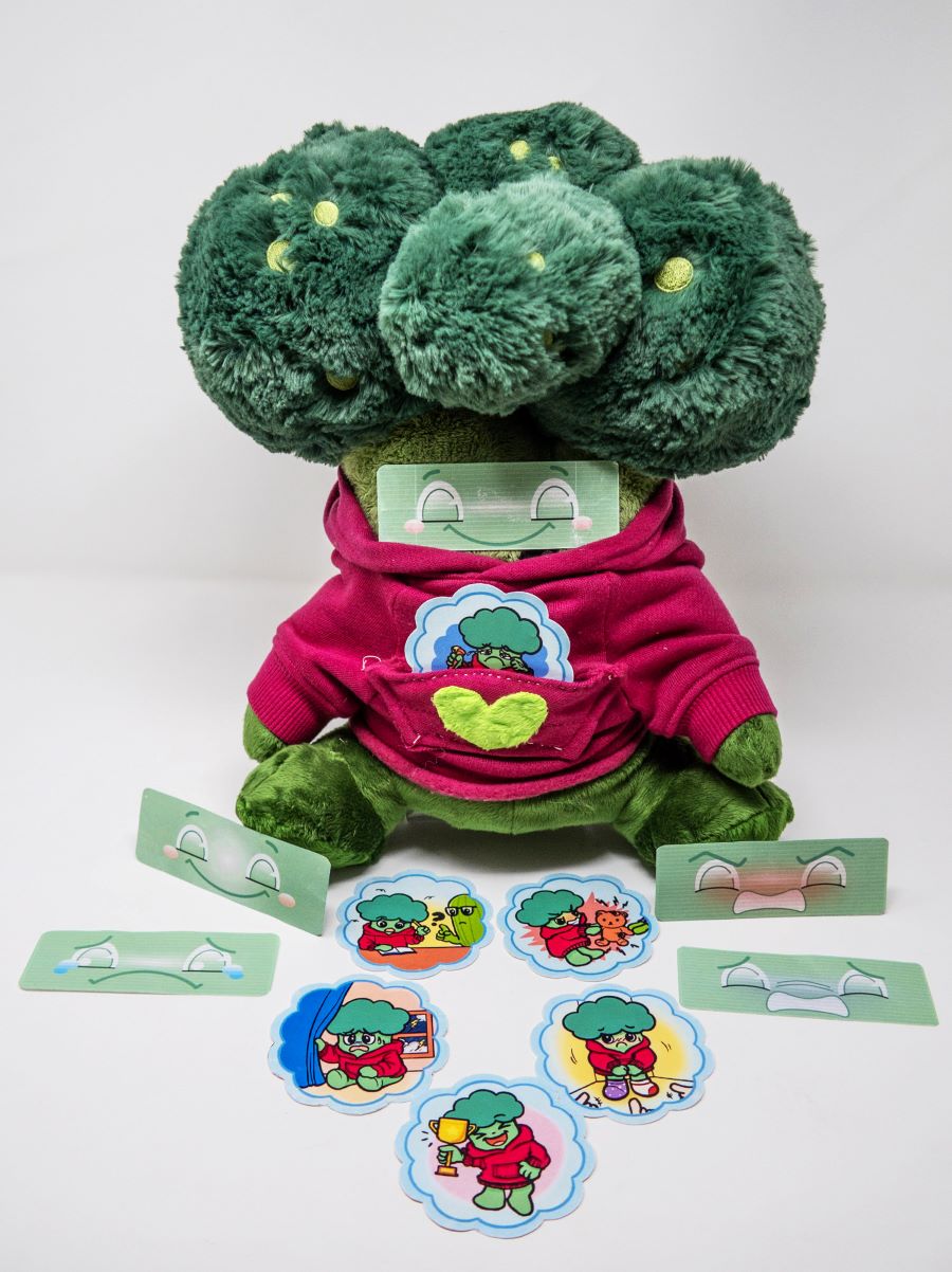 Plush-toy shaped like a broccoli wearing a pink hoodie, displayed with interchangeable illustrated cards showing different activities and facial emotions. The toy is designed as an interactive aid for children to explore feelings and communication.