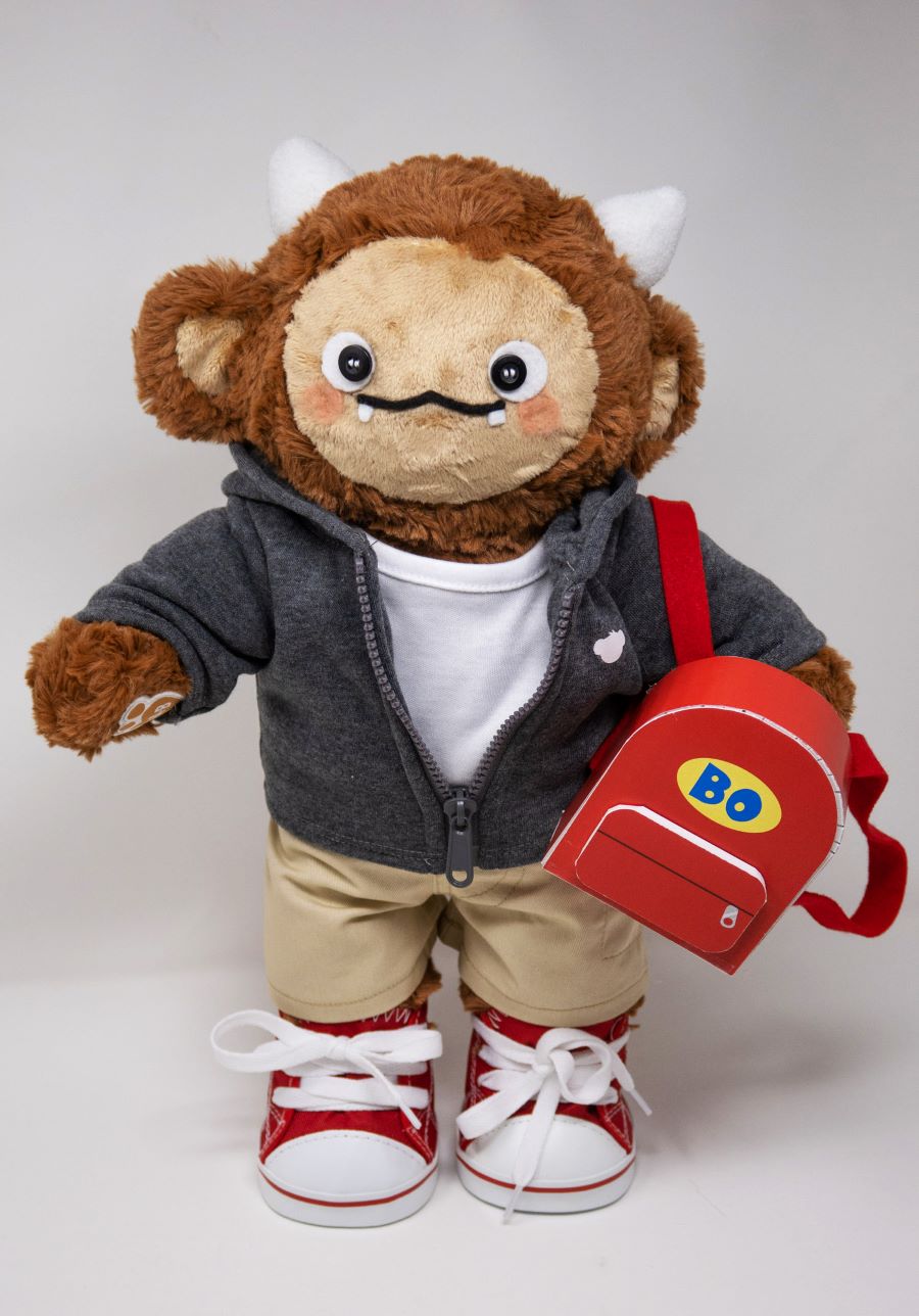 Plush monster doll with brown fur, small horns, and a friendly face. It is dressed in a gray hoodie, khaki pants, and red high-top sneakers. The toy carries a red backpack labeled 'Bo,' suggesting it is designed as a character-based childhood learning companion.