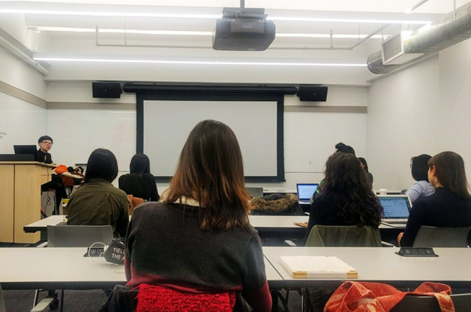 A classroom view from behind students looking toward a instructor and a large projection screen.
