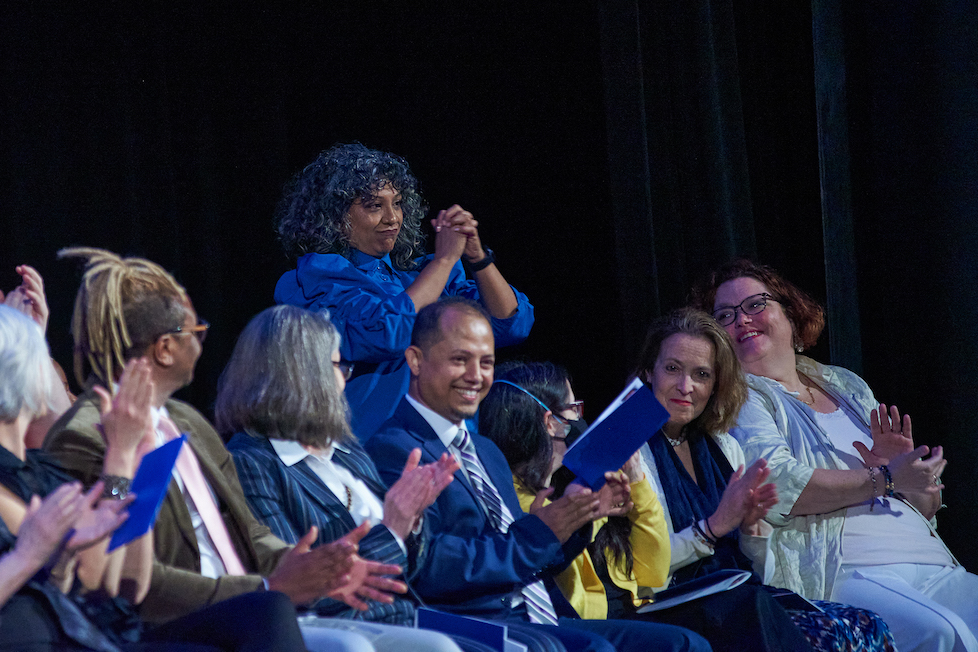 A group of five diverse faculty members smiling and holding award certificates on a stage.