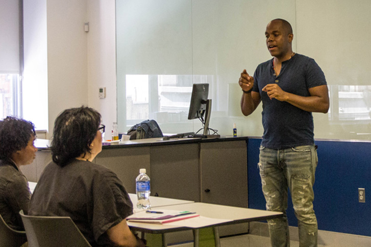 A male instructor standing at the front of a classroom, gesturing while speaking to a seated student.