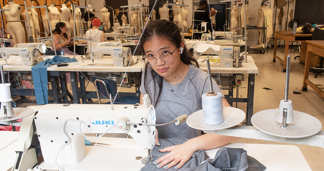  student at a sewing machine in a large classroom with other students working in the background and fitting forms lined allong the walls of the room